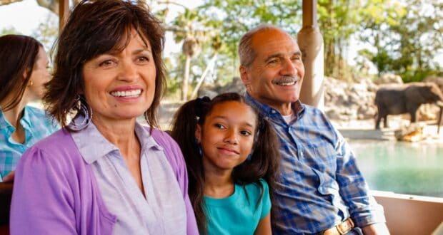 A smiling family is sitting on a Kilimanjaro Safaris ride, with a young girl nestled between two adults. They are enjoying the stunning view, while an elephant meanders in the sunlit background. The scene is bright and cheerful.