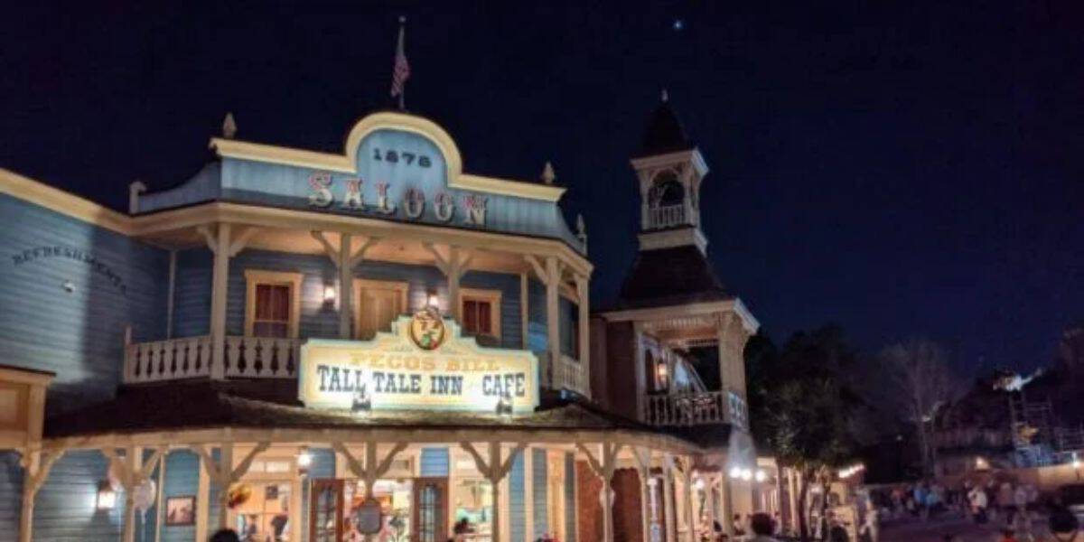 A nighttime photo of a western-themed building with a sign that reads "Pecos Bill Tall Tale Inn and Cafe" and "Saloon." The building has a rustic facade and an old-fashioned clock tower. People are walking around the area. The sky is dark with a few visible stars.