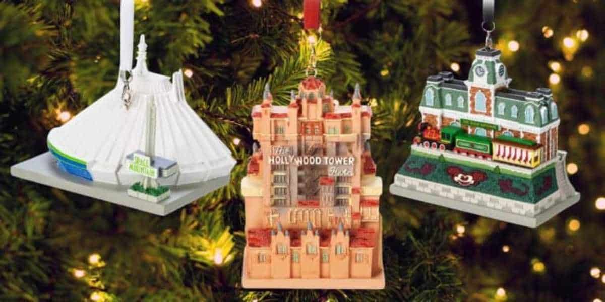 Three theme park ride ornaments hang on a Christmas tree. They depict a futuristic white structure, The Hollywood Tower Hotel, and a train station with a green train. The tree is decorated with warm white lights.
