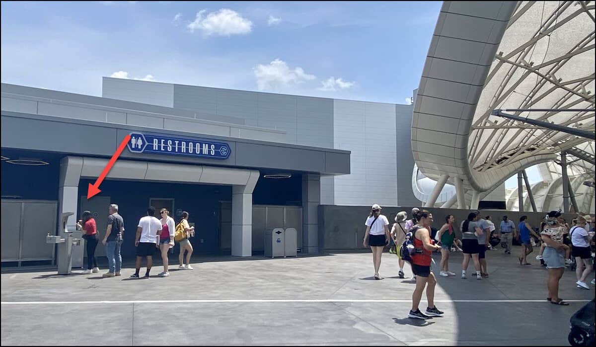 A modern public restroom building under a clear sky, located near a water refill station. Several people are standing nearby, some entering; a red arrow points to a person near the entrance in a red shirt. Others stroll or stand around, enjoying the bright day reminiscent of Magic Kingdom's charm.