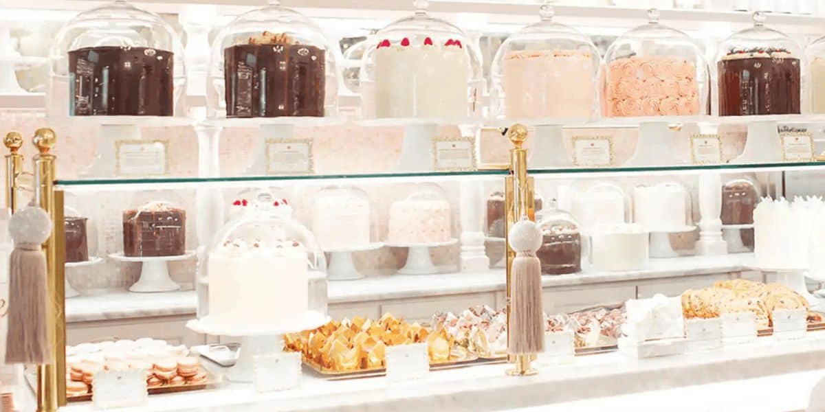 A display case filled with various elegant cakes and pastries, each covered by glass domes. The cakes are arranged on white stands, featuring different flavors and frosting styles, in a bright and inviting bakery setting.