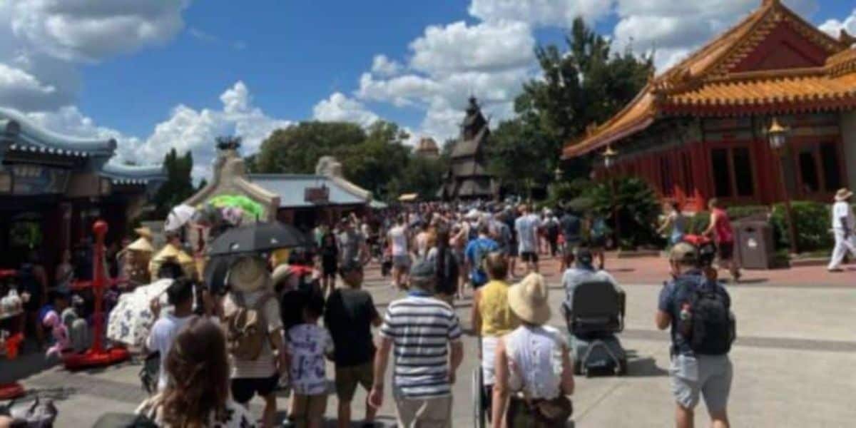 A large crowd of people walk down a wide path in a park or festival setting. Traditional architectural structures with ornate roofs line the walkway, and the sky is partly cloudy.
