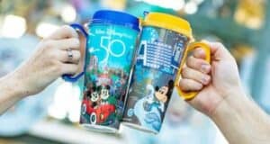 Two hands holding colorful Disney-themed mugs, each adorned with images of Mickey Mouse and Disney landmarks, against a blurred background. The mugs bear "Walt Disney World 50" in celebration of the park's anniversary.