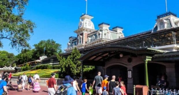 People walk toward the entrance of a theme park on a sunny day. The building features a clock tower and intricate architectural details, with a well-maintained garden in front. Trees and a clear blue sky are in the background.