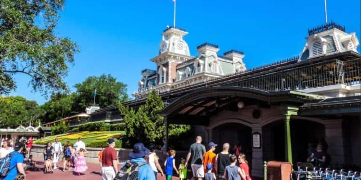 People walk toward the entrance of a theme park on a sunny day. The building features a clock tower and intricate architectural details, with a well-maintained garden in front. Trees and a clear blue sky are in the background.