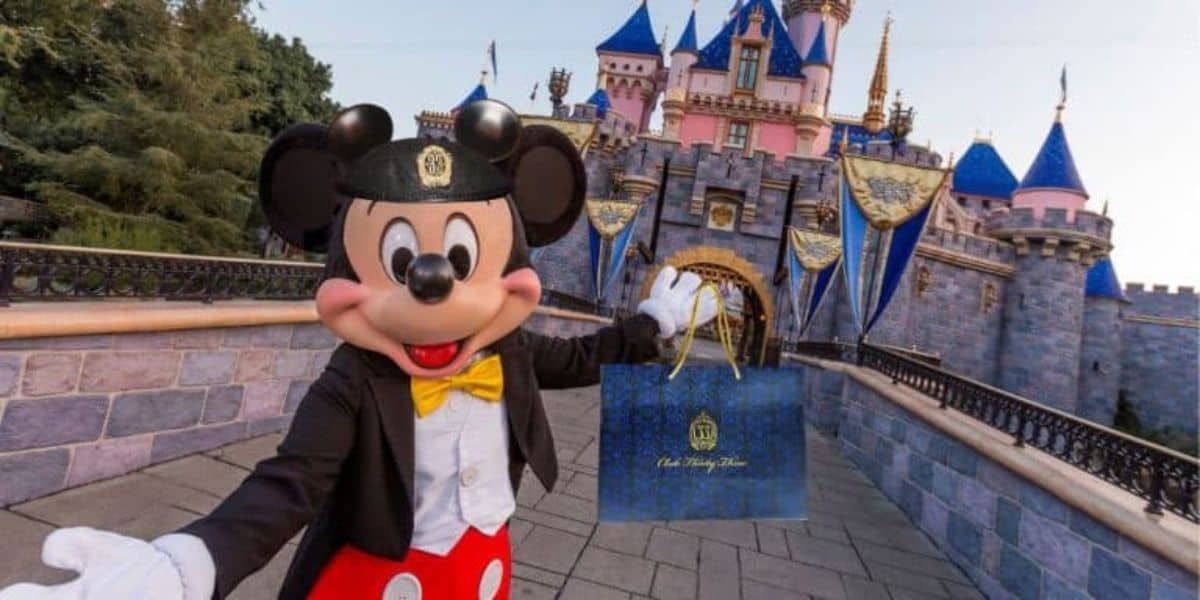 Mickey Mouse, dressed in a tuxedo and hat, stands in front of a pink and blue castle, holding a blue gift bag with a gold design. The setting is a theme park with decorative flags adorning the castle.