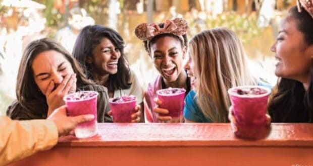 Five women are laughing and holding vibrant purple drinks. They are in an outdoor setting, possibly at a theme park, with blurred greenery in the background. One wears a headband with mouse ears, adding to the joyful atmosphere.