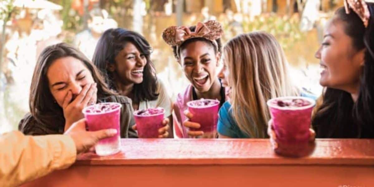 Five women are laughing and holding vibrant purple drinks. They are in an outdoor setting, possibly at a theme park, with blurred greenery in the background. One wears a headband with mouse ears, adding to the joyful atmosphere.