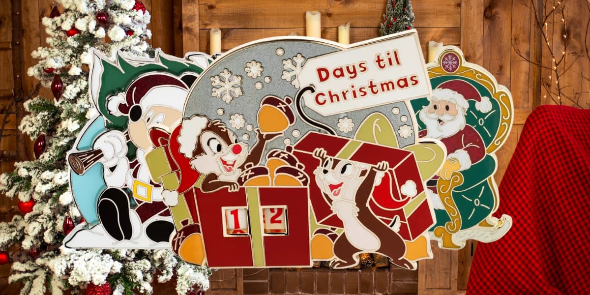 Christmas countdown sign with cartoon reindeer and elves, set against a festive wooden background. The sign reads "Days 'til Christmas" and has numbered blocks displaying "12." Decorated Christmas tree and red plaid chair in the background.