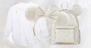 A soft-focus image featuring a white long-sleeve shirt with a subtle design, a white headband with mouse ears and a large bow, and a shiny silver backpack with mouse ears, displayed against a white background.