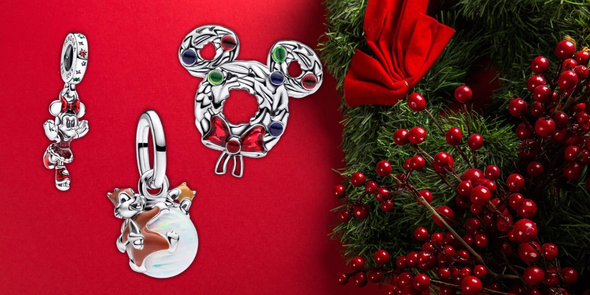 A festive display featuring three Disney-themed charms on a red background. To the right, green foliage and red berries add a holiday touch. The charms include a Mickey Mouse wreath, a character in a Christmas outfit, and a snow globe.