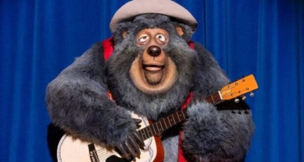 A large anthropomorphic bear wearing a hat and a red vest is joyfully playing a guitar on a stage with a blue curtain backdrop. The bear appears to be part of an animated or animatronic performance.