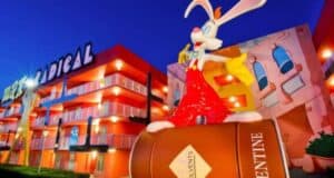 A large cartoon figure of a white rabbit with long ears and wearing red overalls and yellow gloves sits atop a barrel-like structure in front of a brightly colored, multi-story building with balconies. The building features bold, whimsical designs and colors.