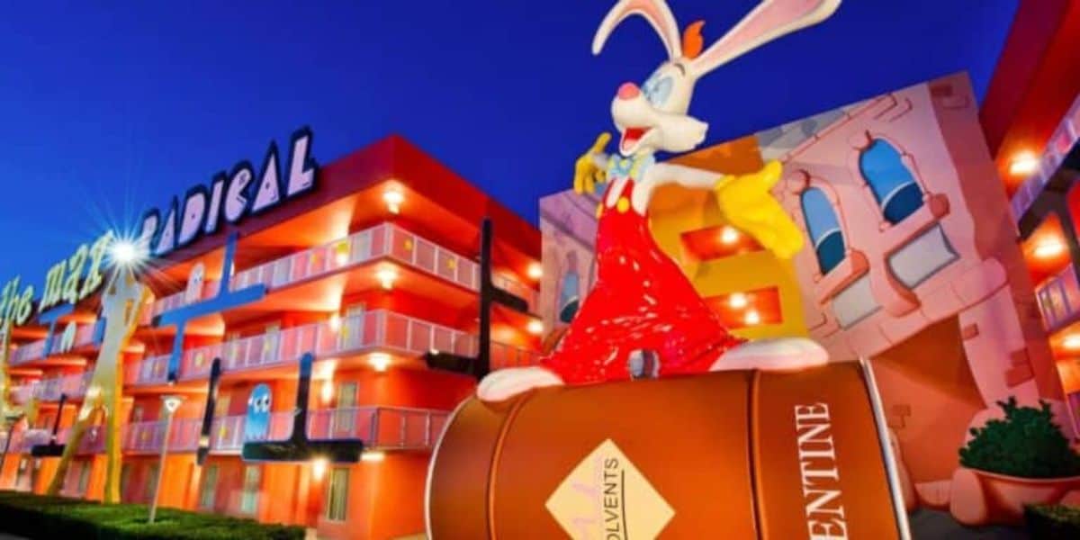 A large cartoon figure of a white rabbit with long ears and wearing red overalls and yellow gloves sits atop a barrel-like structure in front of a brightly colored, multi-story building with balconies. The building features bold, whimsical designs and colors.