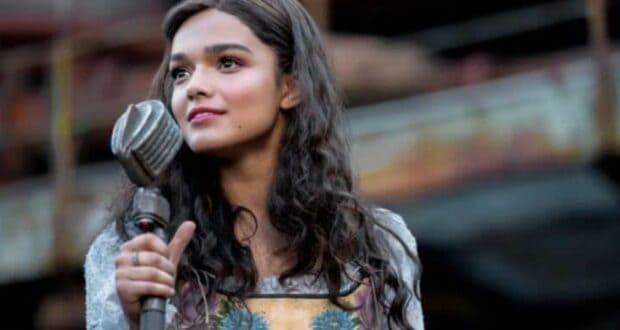 A woman with long, wavy hair stands outdoors holding a vintage microphone. She is wearing a dress with floral embroidery and appears to be singing or speaking, with a thoughtful expression. The background is blurred, showing a rustic setting.