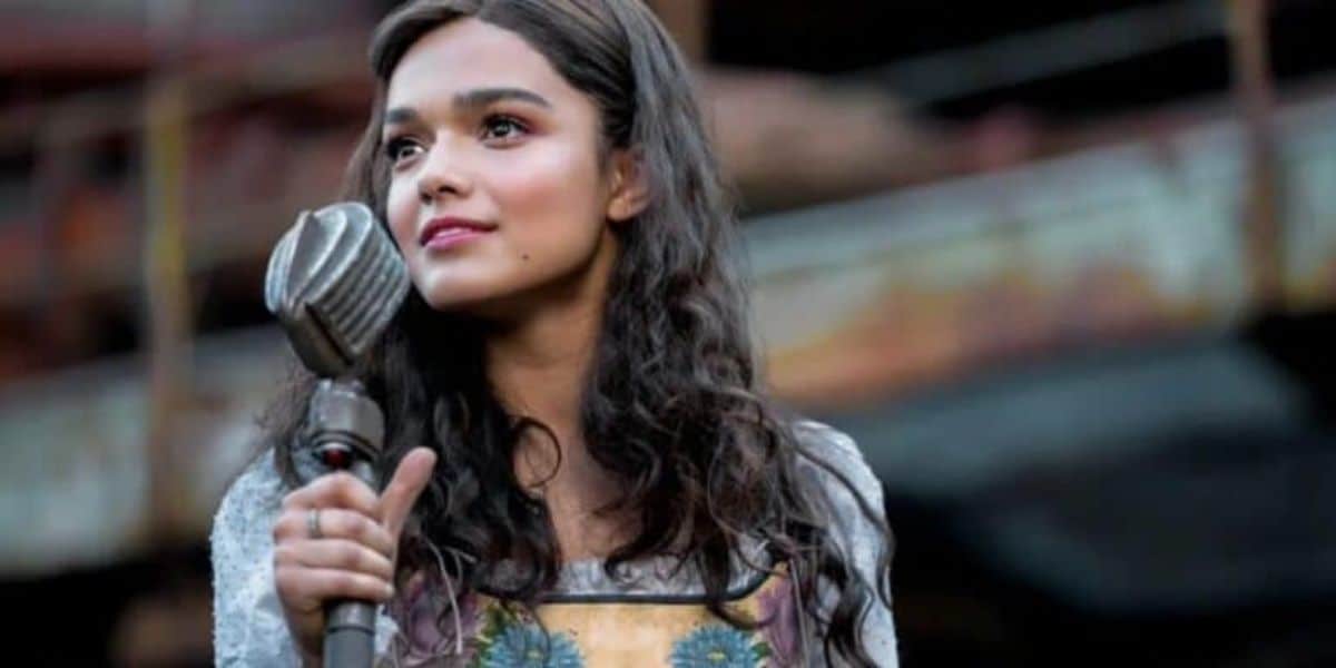 A woman with long, wavy hair stands outdoors holding a vintage microphone. She is wearing a dress with floral embroidery and appears to be singing or speaking, with a thoughtful expression. The background is blurred, showing a rustic setting.