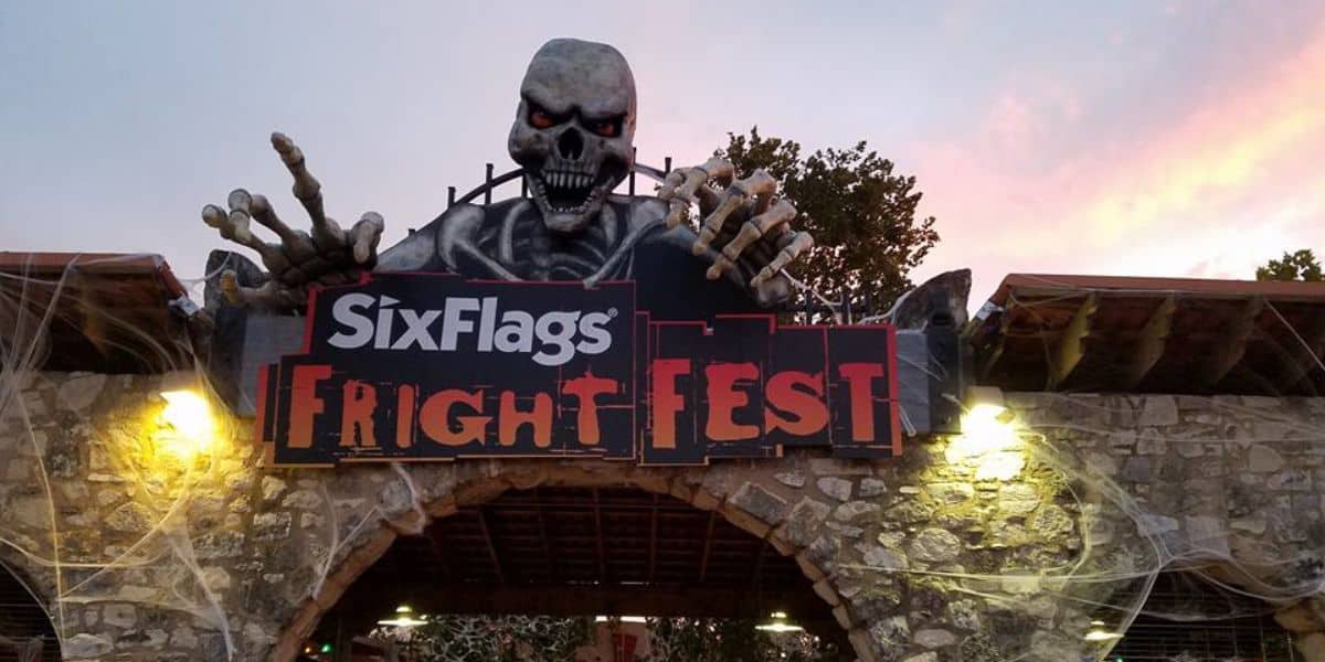Entrance to Six Flags Fright Fest, featuring a large skeleton with red eyes and outstretched hands above a stone archway. The sky is tinged with pink and orange hues, adding to the eerie atmosphere.