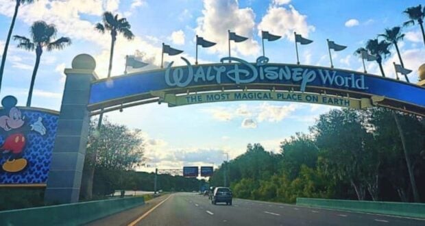 Entrance sign to Walt Disney World with the text "The Most Magical Place on Earth" displayed prominently. A Mickey Mouse image is on the left side of the sign. The road leads past the entrance with trees on both sides and blue sky with clouds overhead.