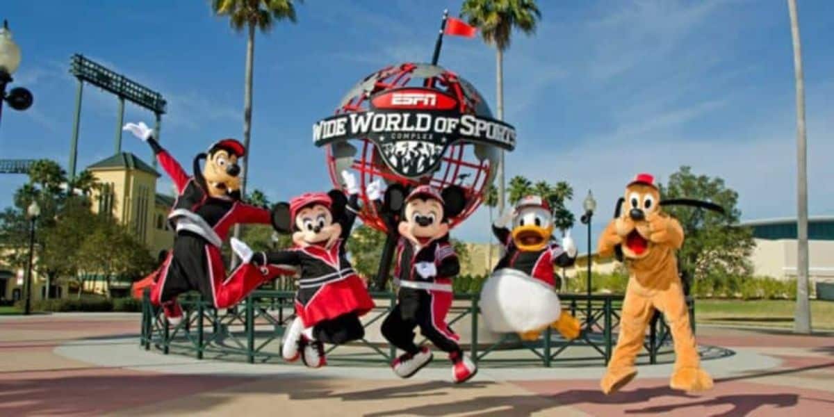 Characters in sports outfits jump in front of the ESPN Wide World of Sports Complex sign. The scene is lively, with palm trees and a clear sky in the background.
