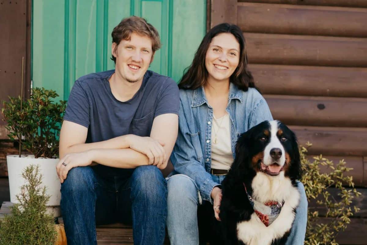 A man and woman sit on wooden steps in front of a log cabin with a green door with their dog.