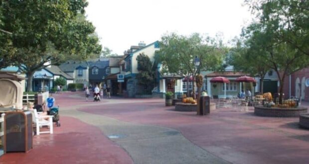 A quiet, sunlit street in a theme park, featuring quaint, colorful buildings, trees, and benches. Few people are strolling, and there's a seating area with tables and red umbrellas on the right side.