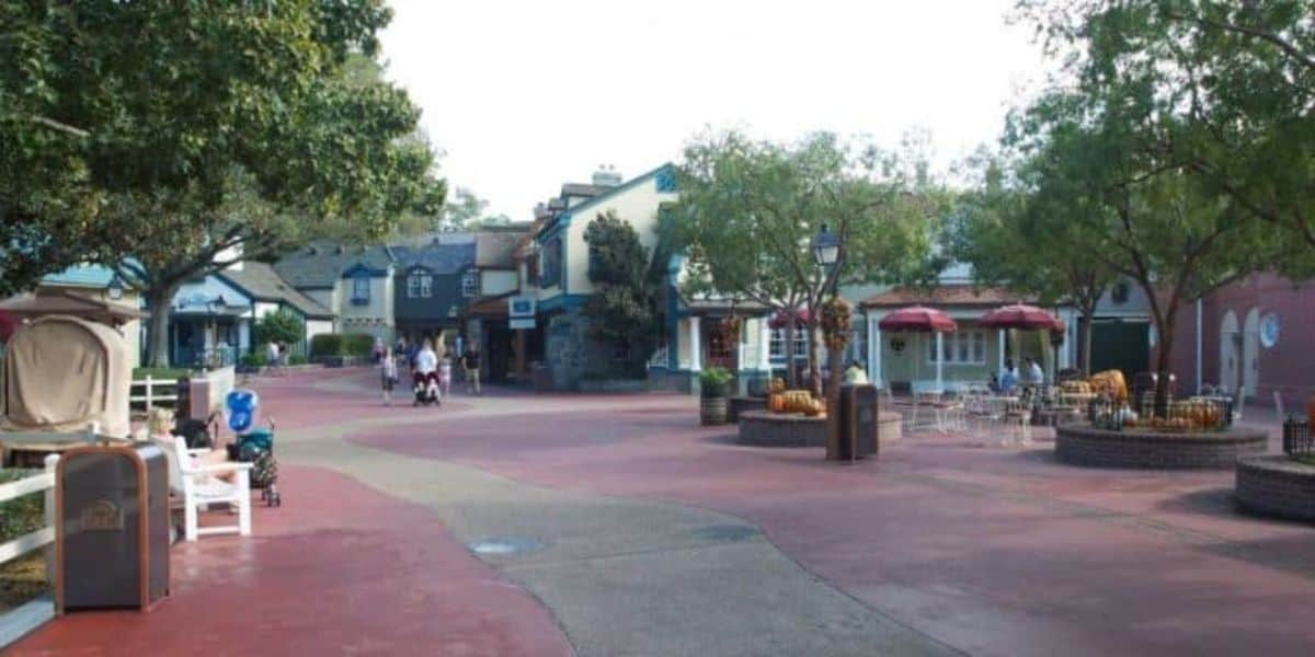A quiet, sunlit street in a theme park, featuring quaint, colorful buildings, trees, and benches. Few people are strolling, and there's a seating area with tables and red umbrellas on the right side.