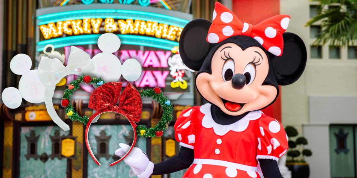 A person in a Minnie Mouse costume stands smiling in front of a colorful marquee. They are wearing a red and white polka-dot dress and bow. In the foreground, festive mouse ear headbands are displayed.