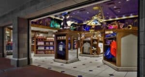 The image shows a brightly lit retail store with various themed merchandise. Shelves and racks display clothing, plush toys, and other items. The decor features magical motifs, including a prominent golden wand and musical notes on the walls and ceiling.