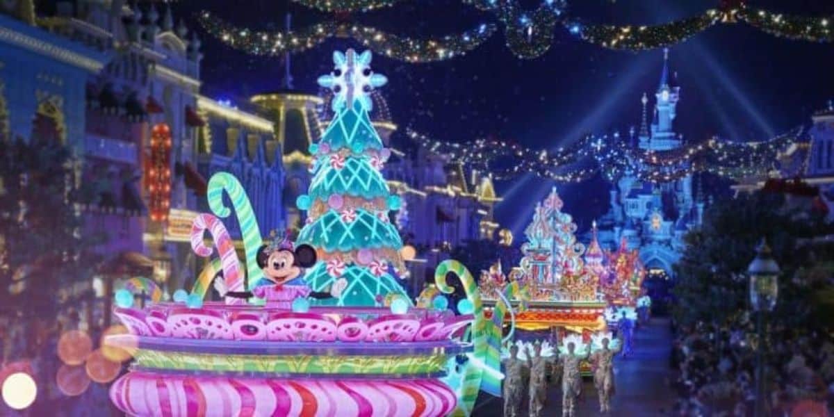 A festive parade at night features brightly lit floats with colorful decorations, including a Christmas tree and candy motifs. A character with large ears and a polka dot bow is on a float. String lights adorn the buildings in the background.