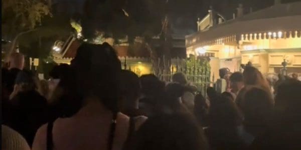 A large crowd of people gathers outside a building at night. The area is dimly lit, with trees and a fence visible in the background. The atmosphere seems lively and bustling.