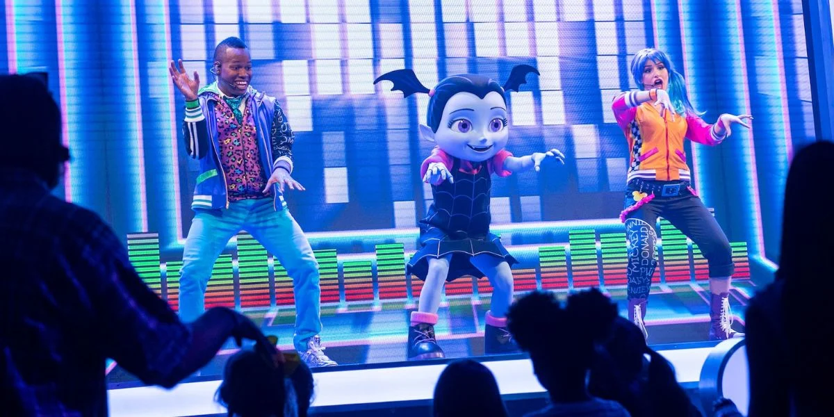 A vibrant stage performance featuring a large, animated vampire bat character in the center flanked by two energetic dancers. The background is colorful with digital graphics, and the audience, mostly children, watches excitedly.