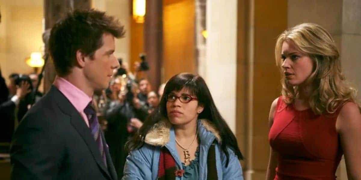 A man in a suit and pink shirt talks to a woman with glasses and braces, wearing a blue coat with a colorful scarf. Another woman in a red dress looks on. They are indoors with people and cameras in the background.
