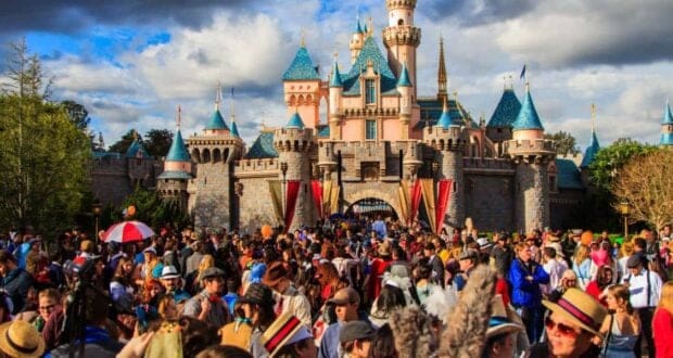 A large crowd gathers in front of a fairy-tale castle with blue spires and pink walls, set against a partly cloudy sky. The scene is lively, with people enjoying a sunny day, some wearing hats and holding balloons.