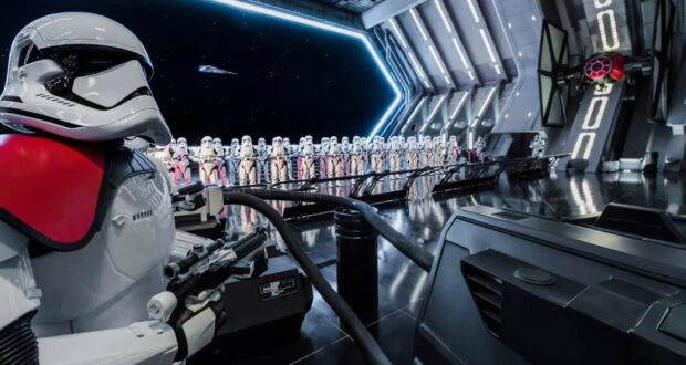 A row of stormtroopers stands inside a futuristic spaceship hangar. A stormtrooper in the foreground has red markings on its armor. The room is metallic and sleek, with a large open bay showing space and a starship.