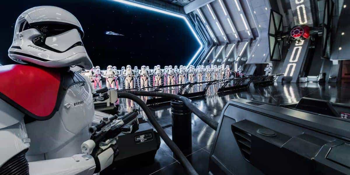 A row of stormtroopers stands inside a futuristic spaceship hangar. A stormtrooper in the foreground has red markings on its armor. The room is metallic and sleek, with a large open bay showing space and a starship.