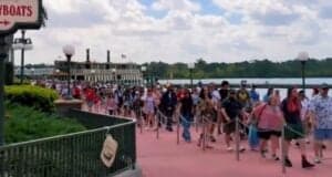A large crowd of people is walking along a pathway near a dock with a ferryboat in the background. The sky is partly cloudy, and trees line the horizon. A sign for "Ferryboats" is visible on the left.