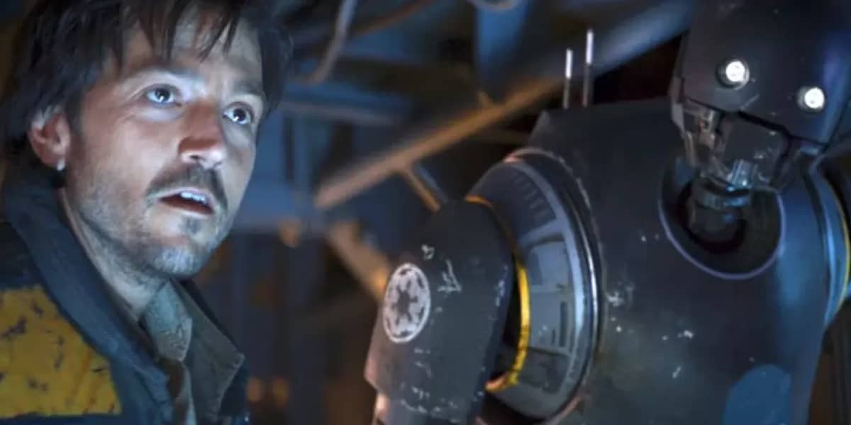 A man with messy hair and a scruffy beard stands next to a tall, black droid with glowing eyes. Both appear tense and are lit by blue and yellow light inside a metallic, industrial setting.