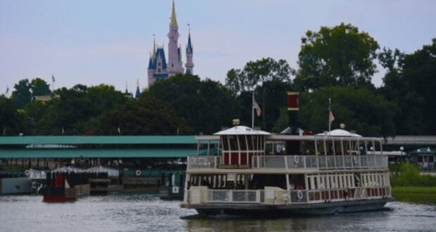 A large white and red ferry boat crosses a calm body of water, with a green-roofed dock and a fairytale castle with blue spires visible in the background among trees.