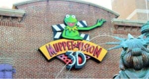 A colorful sign featuring Kermit the Frog is displayed on a brick wall, reading "Muppet*Vision 3D." In the foreground, part of a statue resembling the Statue of Liberty is visible.