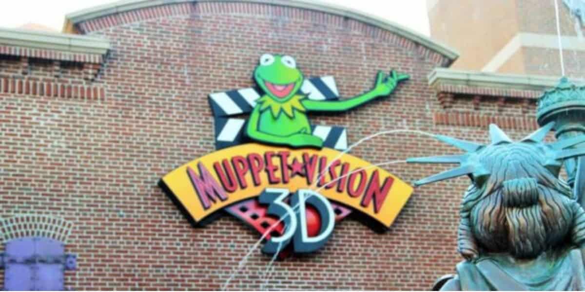 A colorful sign featuring Kermit the Frog is displayed on a brick wall, reading "Muppet*Vision 3D." In the foreground, part of a statue resembling the Statue of Liberty is visible.