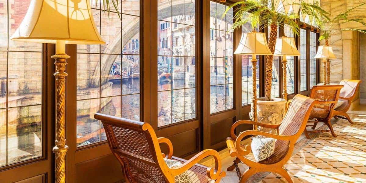 Sunny indoor seating area with elegant wooden chairs, patterned cushions, tall lamps, potted palm plants, and large windows offering a view of a picturesque canal and arched stone bridge outside.
