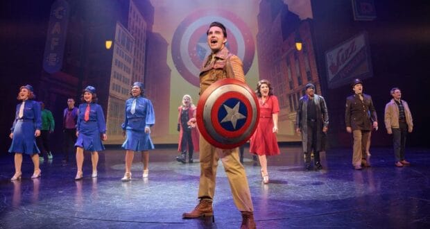 Group of performers in retro costumes sing on stage. One holds a large star-emblazoned shield. A cityscape backdrop adds depth to the scene, with vintage signage visible. The atmosphere is lively and theatrical.