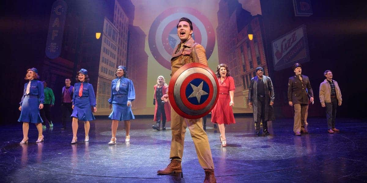 Group of performers in retro costumes sing on stage. One holds a large star-emblazoned shield. A cityscape backdrop adds depth to the scene, with vintage signage visible. The atmosphere is lively and theatrical.
