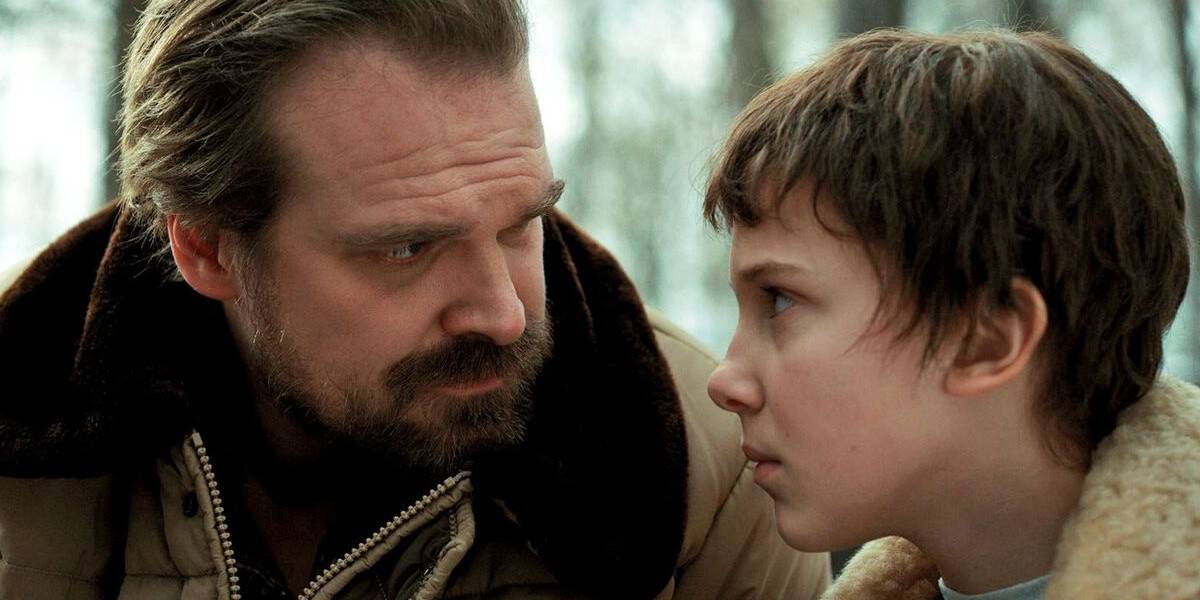 A bearded man in a brown jacket looks seriously at a young person with short hair and a fur-collared coat; both appear to be outdoors in a tense moment.