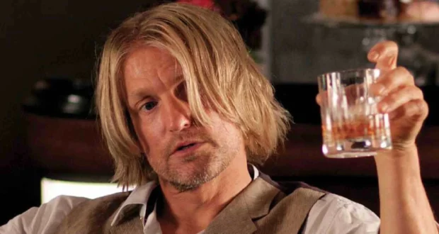 A man with shoulder-length blond hair and a beard holds up a glass of brown liquor, looking slightly disheveled and contemplative. He is wearing a beige vest over a white shirt.
