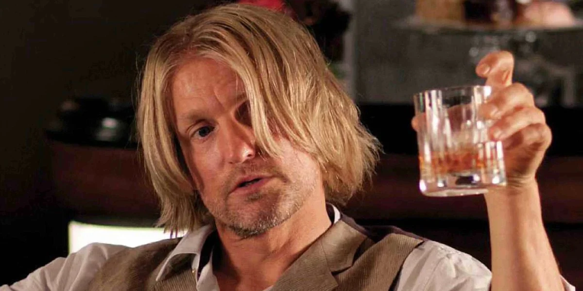A man with shoulder-length blond hair and a beard holds up a glass of brown liquor, looking slightly disheveled and contemplative. He is wearing a beige vest over a white shirt.