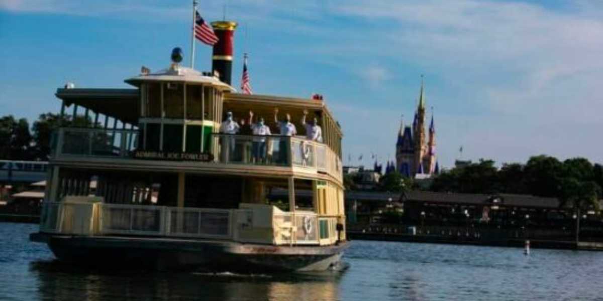 A ferryboat named Admiral Joe Fowler crosses a lake with several people on board; American flags are flying, and a castle is visible in the background under a blue sky.