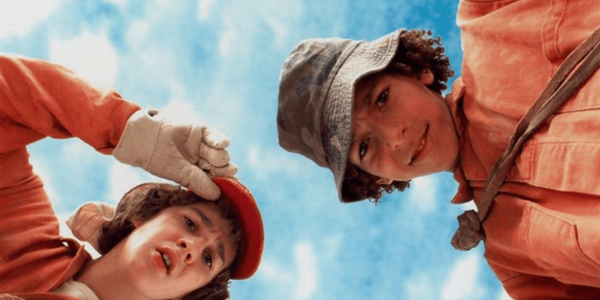 Two teens in orange uniforms and hats look down toward the camera against a blue sky, appearing curious or puzzled. One wears gloves and adjusts their red cap, while the other wears a bucket hat.