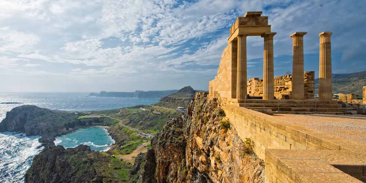 Ancient stone columns of the Lindos Acropolis stand on a cliff overlooking a rugged coastline, blue sea, and a small beach below, under a partly cloudy sky in Rhodes, Greece.