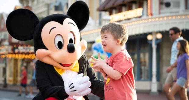 A young boy excitedly interacts with a person in a Mickey Mouse costume at an amusement park, with colorful buildings and people in the background.